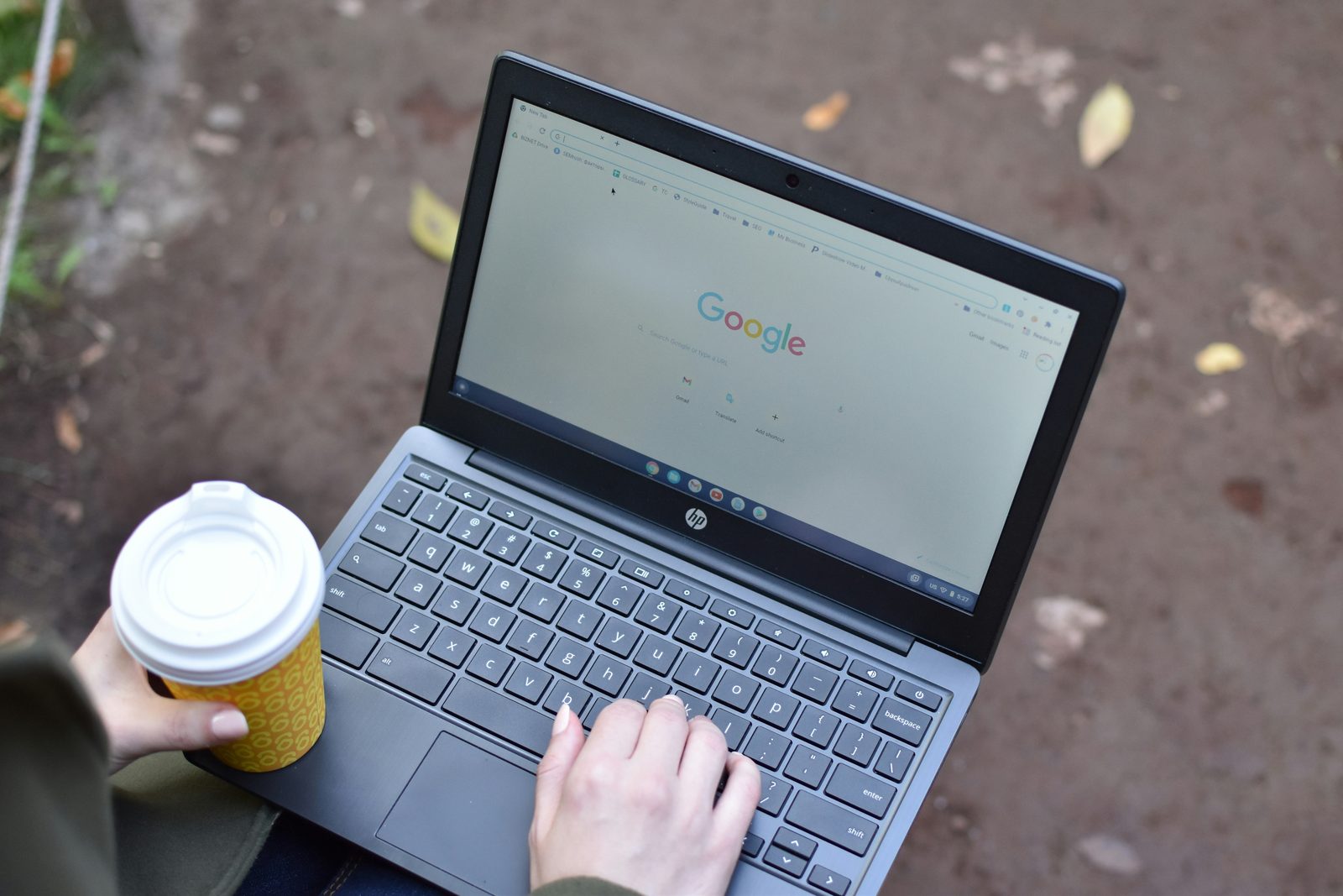 Laptop op schoot met de startpagina van Google op het scherm, buiten, met een koffiebeker in de hand