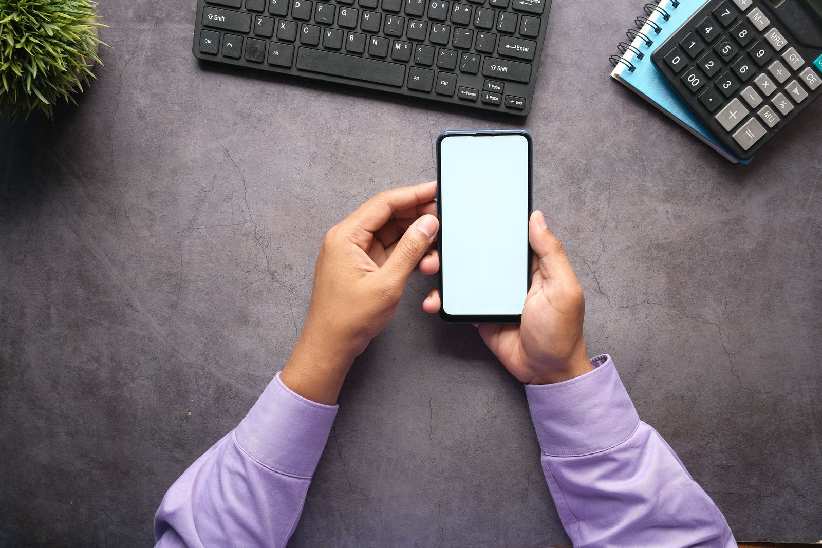 Twee handen houden een telefoon met een leeg scherm boven een bureau met een toetsenbord en een rekenmachine