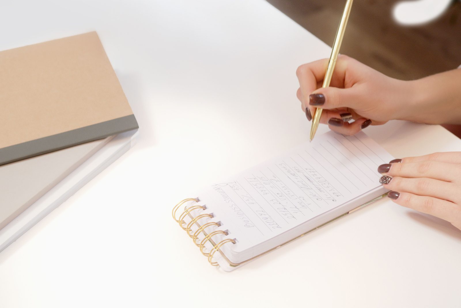 Twee handen die met een potlood in een klein notitieboekje schrijven op een licht bureau